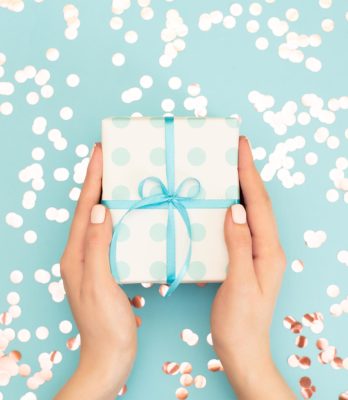 Womans hands holding gift or present box decorated confetti on pink pastel table top view. Flat lay composition for birthday or wedding