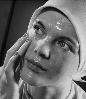 A black and white photo of a woman rubbing a moist, shiny beauty care product on her face. (Photo by George Karger/Getty Images)